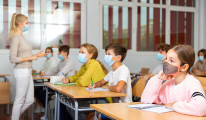 Smart teenager in protective mask studying in classroom, listening to lecturer and writing in...