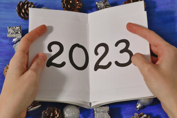 hands holding the inscription 2023 on paper and below which are silver Christmas decorations and gifts and pine cones on a dark blue background