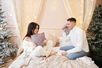 Fototapeta premium Happy lovely couple having fun, fighting with pillows on bed in New Years festively decorated bedroom, two teenagers enjoying company of each other, spending time at home during Christmas