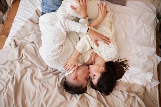 Top view of passionate romantic couple spending time before New Year at home with garlands. Sexy young woman and handsome man are lying on bed in sweaters and hugging.
