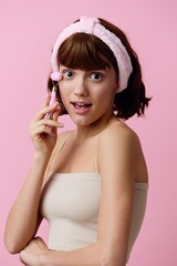 vertical photo of cute, elegant, happy woman doing a facial massage with a pink roller, standing on a pink background, with a soft headband on her head. Beauty, skin care and youth maintenance topics
