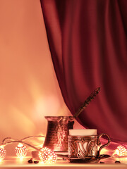 Turkish coffee pot and traditional cup with oriental sweets. Ornate copper metal garland and tea lights. Vivid red and golden monochromatic festive background lit by candlelight.