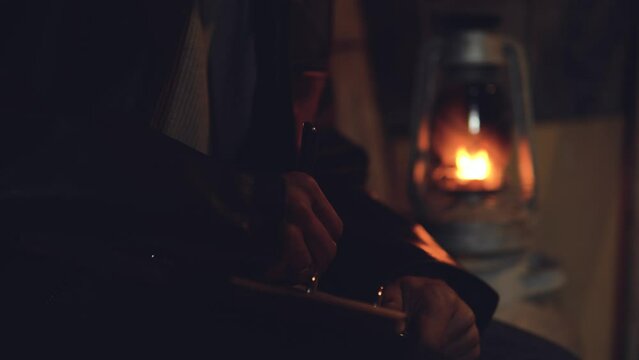 Adult seriously man with beard write old diary wear a vintage leather jacket in retro small fishing house of wood at deep night. Vintage a kerosene lamp fire