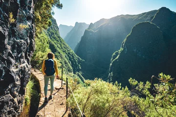 Fotobehang Backpacker woman walking along sunny hiking trail below large rock wall along water channel at steep cliff through Madeira's rainforest. Levada of Caldeirão Verde, Madeira Island, Portugal, Europe. © Michael