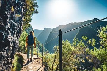 Backpacker woman walking along sunny hiking trail below large rock wall along water channel at...