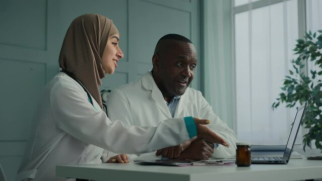 Medical people two professional doctors colleagues arabian young woman therapist discuss electronic health test results with african senior man use computer application doing online digital diagnostic