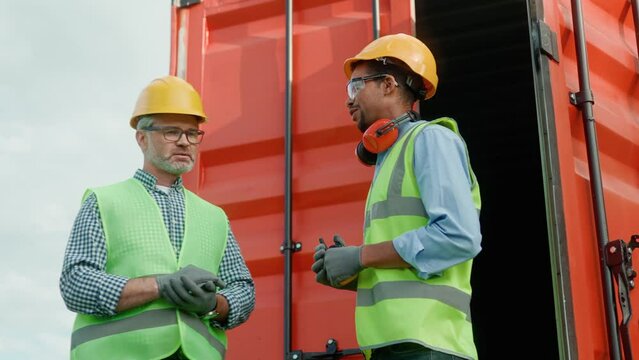 Side-view of two hard-working colleagues working on cargo transportation company talking in front of opened container. African-American and caucasian men discussing wearing safety equipment.