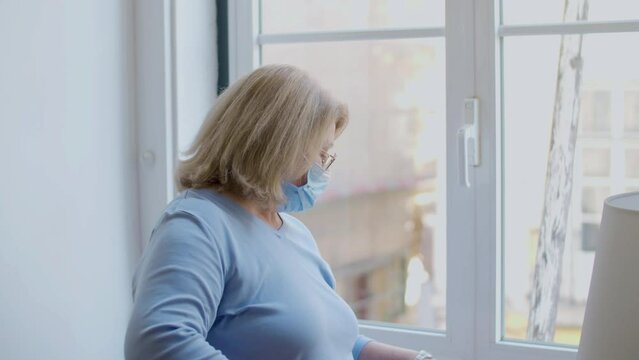 Side view of mature woman in medical mask standing near window. Pensive woman resenting doctor for being late, worrying about vaccination against covid, checking time on wristwatch. Healthcare concept