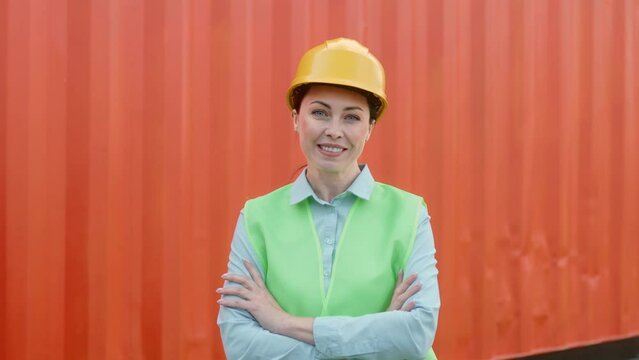 Portrait hard-working happy employee of transportation company standing with crossed arms with red container on background. Cheerful female worker wearing safety equipment looking at camera smiling.