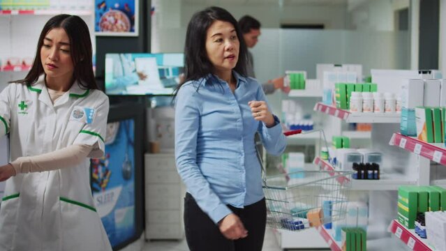 Asian customer asking pharmacist for help with medicaments, sarching for prescription medicine in bottle. Consultant checking supplement and plls boxes to give treatment to person.