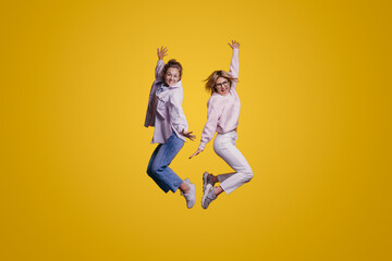 Full body size portrait of two positive adorable good-looking women jumping isolated on yellow background raising hands up. Attractive beautiful girl. Trendy