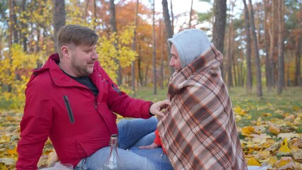 Side view Caucasian gay man talking with partner wrapping in blanket smiling. Happy loving LGBTQ couple sitting on blanket on picnic outdoors on autumn day