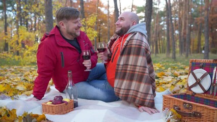Loving gay couple toasting clinking red wine glasses drinking enjoying autumn picnic on sunny day. Wide shot side view portrait of smiling happy LGBT Caucasian men dating outdoors