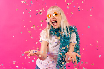 Smiling at a party throwing confetti in sunglasses, blonde caucasian girl on pink background studio