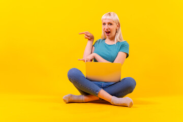 Fototapeta na wymiar Sitting with a laptop pointing to the left, blonde caucasian girl in studio on yellow background