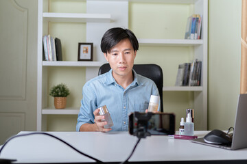 Young adult Asian beauty video blogger man filming, online live streaming his video blog about male cosmetic, male skincare. Guy looking camera to show new skin care for men product, teaching make up.
