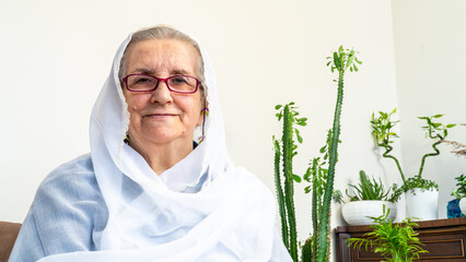 Portrait of old Muslim woman at eighties wearing white hijab sitting at home while fasting during...