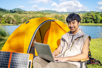 Young Asian traveling man relax and enjoy outdoor lifestyle camping in forest mountain on summer holiday. Attractive male relaxing in chair while working on laptop during camping day in nature