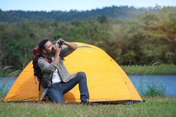 Backpacker man uses binoculars to watch birds while in front of a yellow tent at campsite,...