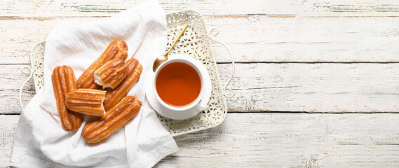 Tray with sweet tasty eclairs and hot tea on light wooden background with space for text, top view
