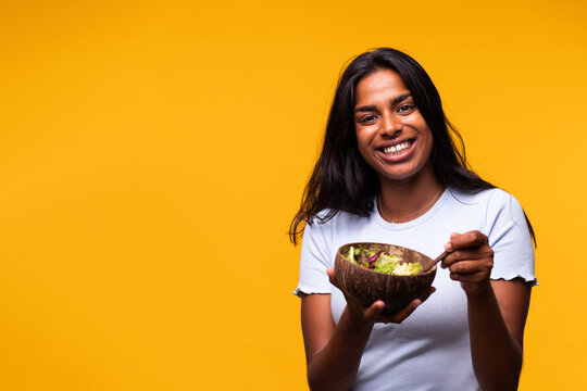 Happy Young Indian Woman Eating Healthy Salad Looking At Camera. Isolated On Yellow Background. Studio Shot.