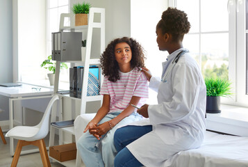 Friendly female pediatrician touches shoulder of teenage girl and informs her that there is no threat to her health. African American doctor and her preteen patient are talking in hospital office.