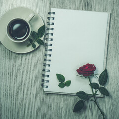 An open notebook with white sheets. An office desk with a notepad, a red rose and a cup of coffee. Flat layout, top view, place to copy. Space for text. Selective focus.