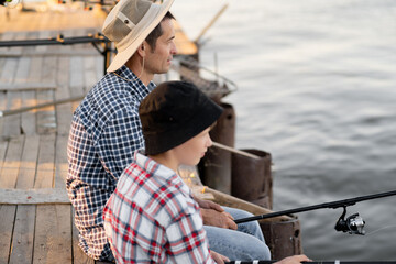 Side view of father and son sitting together on pier fishing with rods in calm lake waters with landscape of setting sun, both wearing checkered shirts.