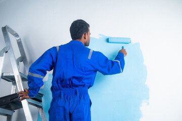 Back view shot of painter doing painting work on wall with roller while standing on ladder - concept of - concept of hard working, blue collar job and home renovation.
