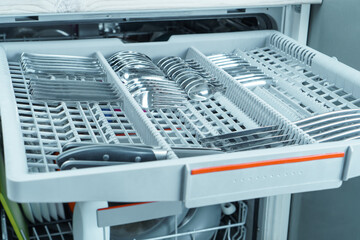Close-up of grey compartment tray of open dishwasher machine with cutlery set of knives, spoons,...