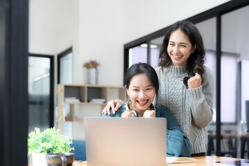 Two young Asian women show joyful expression of success at work smiling happily with a laptop computer in a modern office.