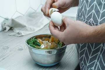Cropped photo of man wearing apron peeling hardboiled egg over metal bowl full of carrot peelings,...