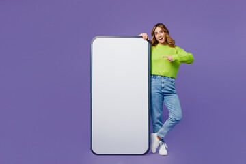 Full body young smiling fun woman 30s she wear casual green knitted sweater stand point finger on big huge blank screen mobile cell phone with area isolated on plain pastel purple background studio.