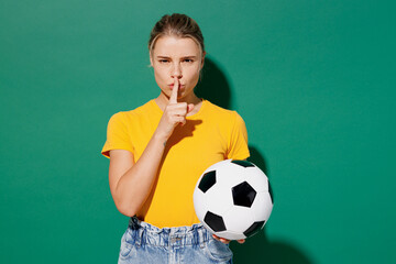 Young woman fan wears basic yellow t-shirt cheer up support football sport team hold in hand soccer ball watch tv live stream say hush shhh be quiet finger on lips isolated on dark green background.