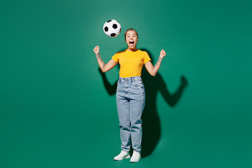 Full body overjoyed young woman fan wear basic yellow t-shirt cheer up support football toss up soccer ball watch tv live stream do winner gesture look camera isolated on dark green background studio.