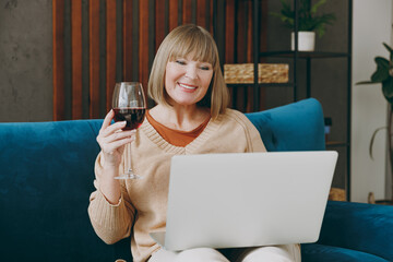 Elderly woman 50s years old wear casual clothes sits on blue sofa use laptop pc computer watch movie film drink wine stay at home flat rest relax spend free spare time in living room indoor grey wall