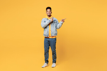 Full body young fun man of African American ethnicity wear denim jacket hoody point index finger aside on area indicate on workspace area copy space mock up isolated on plain yellow background studio.