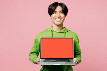 Young IT man of Asian ethnicity wear green hoody hold use work on laptop pc computer with blank screen workspace area isolated on plain pastel light pink background studio. People lifestyle concept.