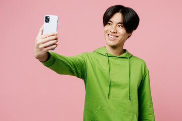 Young fun man of Asian ethnicity wear green hoody doing selfie shot on mobile cell phone post photo on social network isolated on plain pastel light pink background studio. People lifestyle concept.