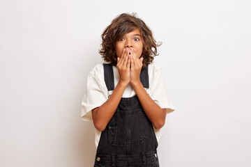 Studio shot of shocked emotional young guy covers mouth with hands, tries to be silent, poses over white background