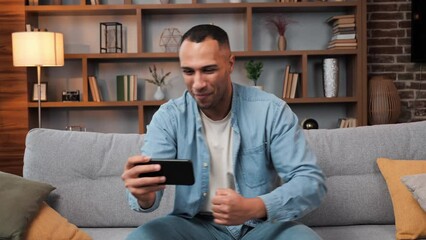 Attractive excited dark skinned guy cheers for favorite football team in mobile phone watching over sports betting at home African american man watch tv sport game celebrate goal team victory on sofa