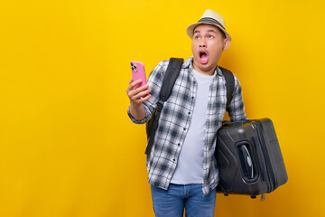 Worried young traveler tourist Asian man 20 years old wears casual clothes hat with backpack holding suitcase and mobile phone, looking aside isolated on yellow background. Air flight journey concept