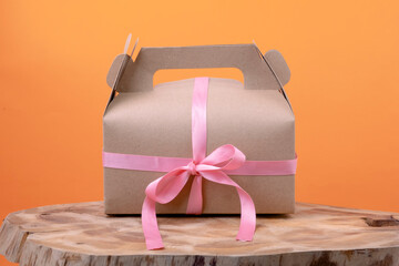 Craft a gift box with a small pink ribbon on wood on orange background. Eco-friendly New Year and Christmas decoration concept