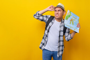 Confused young traveler tourist Asian man wearing casual clothes and hat with a backpack holding city map and put hand on his head, looking aside on yellow background. Air flight journey concept