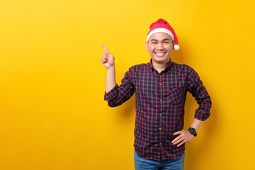 Cheerful young Asian man in Santa hat pointing fingers aside at blank space for advertising text on yellow studio background. Happy New Year 2023 celebration merry holiday concept