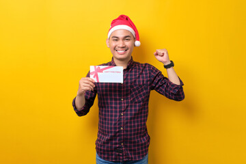 Excited young Asian man in Santa hat holding gift certificate, doing winner gesture over yellow studio background. Happy New Year 2023 celebration merry holiday concept