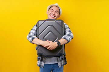 Enjoy traveler tourist Asian man in casual clothes hat backpack keeping his eyes closed hugging a suitcase bag on yellow background. Passenger travel abroad weekends getaway Air flight journey concept - obrazy, fototapety, plakaty