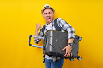 Fototapeta na wymiar Excited traveler tourist Asian man in casual clothes hat backpack hold under hand suitcase bag, clenches his fists and says yes isolated on yellow background