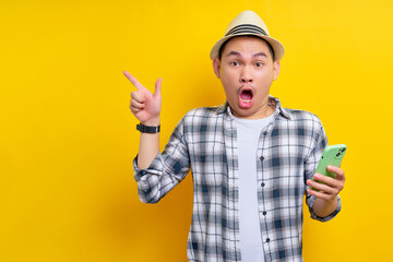 Amazed traveler tourist Asian man in casual clothes hat holding mobile phone and pointing at copy space on yellow background. Passenger traveling on weekends. Air flight journey concept