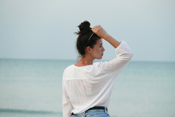 Slim girl walking by the sea. Summer lifestyle image of happy woman walking on the beach. Pretty young woman with long hair  in a white shirt and denim shorts.